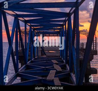 Plan conceptuel de l'homme pêchant sur la jetée abandonnée, pont fait de barres métalliques pendant le coucher du soleil dramatique Banque D'Images