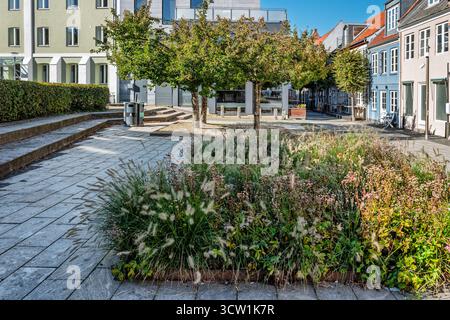 Une scène de rue ensoleillée avec une rangée de tables de café à l'extérieur des vitrines en briques, des plantes en pot et des piétons se promenant le long d'une voie piétonne pavée. A wa Banque D'Images
