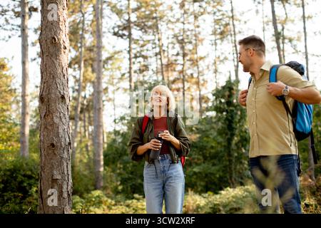 Une femme et un homme font une pause dans une forêt ensoleillée, souriant et bavardant. Ils portent du matériel de randonnée, portant des sacs à dos, entourés de grands arbres et d'une végétation luxuriante Banque D'Images
