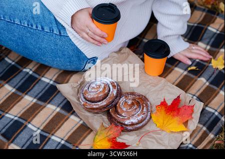 Scène de pique-nique d'automne en gros plan avec vue partielle de la femme en pull assis sur la couverture à carreaux, tenant la tasse à café près des rouleaux de cannelle et l'automne coloré Banque D'Images