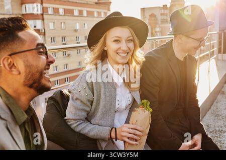 Groupe de jeunes amis élégants assis sur le toit-terrasse avec des collations, bavardant et profitant de la soirée, belle femme blonde souriant à la caméra Banque D'Images