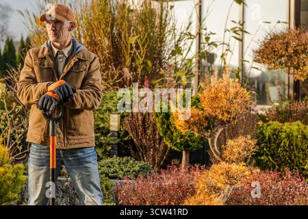 Un homme se tient debout avec une pelle dans une pépinière de plantes, entouré de diverses plantes et arbustes, profitant de la lumière chaude de l'après-midi tout en se préparant au jardinin Banque D'Images