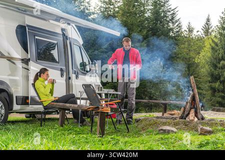 Un homme prépare de la nourriture sur un feu de camp tandis qu'une femme profite d'une boisson chaude assise près d'un camping-car dans un cadre forestier serein. Banque D'Images