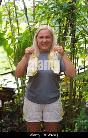 Une femme se tient dans un jardin animé, affichant fièrement deux moitiés de fruits de durian frais. La scène est remplie de plantes luxuriantes et de lumière du soleil, cre Banque D'Images
