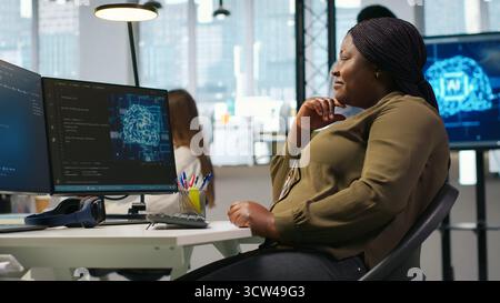 Informaticien dans un bureau de démarrage examinant les métriques du système d'IA, surveillant les tableaux de bord de performance. Femme afro-américaine accordant des réseaux neuronaux en utilisant des outils sur PC, caméra A. Banque D'Images