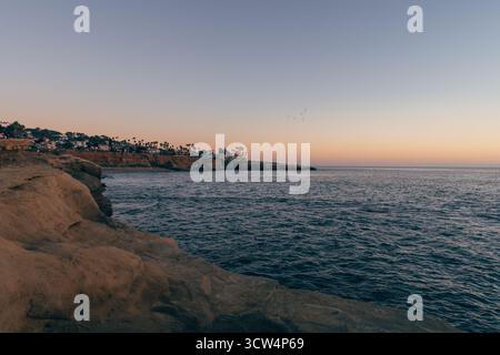 Des couleurs douces remplissent le ciel alors que le soleil se couche sur des eaux calmes. Falaises et palmiers encadrent la vue sereine sur la côte, créant une atmosphère paisible. Banque D'Images
