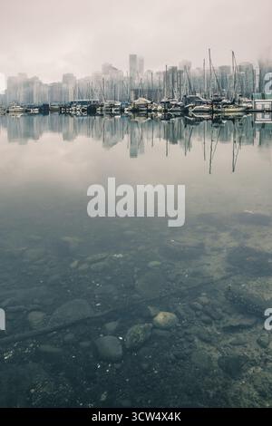 Une scène sereine montre un front de mer brumeux avec des bateaux amarrés reflétant dans des eaux calmes, tandis que la ville se profile en arrière-plan. L'humeur est tranqui Banque D'Images