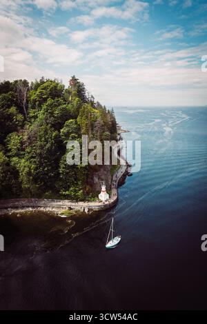 Une belle côte dispose d'un phare debout haut au milieu de la verdure. Un voilier est ancré à proximité sur des eaux calmes sous un ciel dégagé, créant un p Banque D'Images