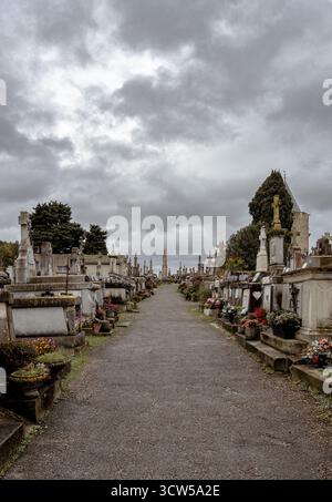 Un petit chemin à travers un cimetière avec des pierres tombales et des pierres tombales avec des croix sous un ciel nuageux. Banque D'Images