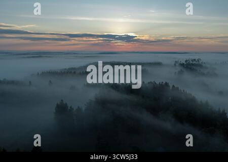 Vue aérienne d'une mer de brouillard couvre un paysage boisé sous un ciel pastel, le soleil regardant à travers les nuages, Raasepori, Uusimaa, Finlande. Banque D'Images