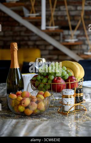 Une bouteille de champagne et un panier de fruits sur une table dressée dans un intérieur moderne. Une table festive avec une corbeille de fruits, une bouteille d'alcool et des épices Banque D'Images