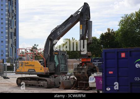 Démolition en cours de l'ancienne gare routière de Gosport, Hampshire, Angleterre. Une pelle Volvo portant la marque « Bath Demolition » domine le site, son bras hydraulique prêt à l’action. À proximité, un conteneur violet et un bac rose ponctuent le sol parsemé de débris. En arrière-plan, un grand bâtiment avec des panneaux bleus et blancs s’élève au-dessus des arbres, contrastant les machines industrielles avec la rénovation urbaine. Banque D'Images