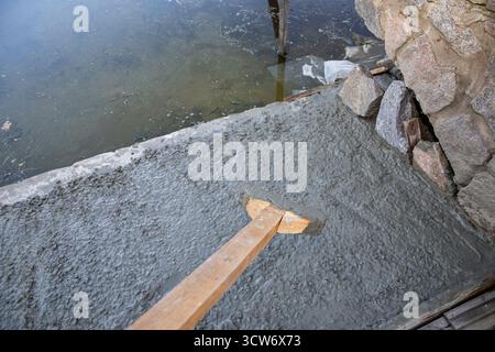 Niveau de l'ouvrier surface de béton fraîchement coulé près du mur de pierre et de l'eau à l'aide d'un outil en bois. Banque D'Images