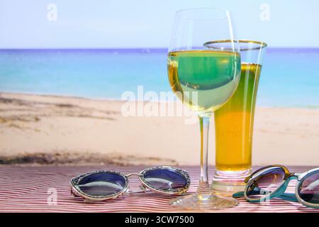 La scène se déroule sur une plage, avec l'océan en arrière-plan. Un verre de vin et un verre de bière sont assis sur une table à côté d'une paire de lunettes de soleil. Scène Banque D'Images