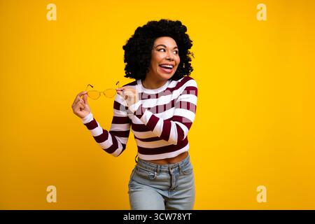 Jeune femme tenant des lunettes et souriant joyeusement sur un fond jaune vif portant un haut rayé Banque D'Images