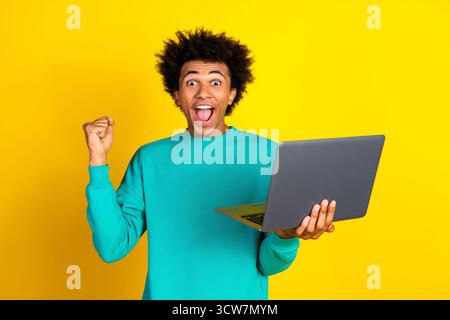 Jeune homme excité célèbre le succès tout en utilisant un moment joyeux d'ordinateur portable sur fond jaune vibrant Banque D'Images