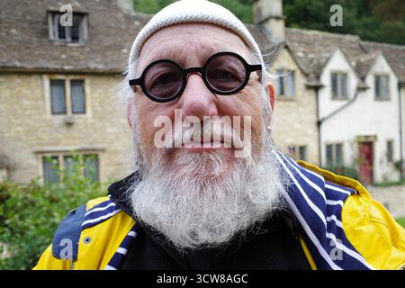 Un homme âgé avec une barbe blanche, des lunettes noires rondes, un bonnet en tricot blanc et un imperméable jaune se tient devant une cotta en pierre traditionnelle de style Cotswold Banque D'Images