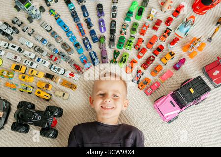 Enfant heureux encadré par des lignes de véhicules miniatures sur tapis, jouets lumineux et expression joyeuse créent une composition ludique Banque D'Images