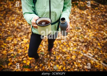 Rafraîchissement avec boisson chaude pendant la randonnée d'automne. Femme touriste buvant du thé de la tasse de voyage dans la forêt Banque D'Images