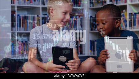 Des garçons souriants d'âge scolaire qui étudient les tableaux financiers sur des tablettes dans le coin de la bibliothèque, avec des étagères Banque D'Images