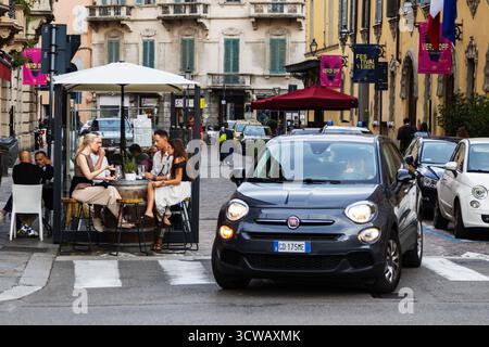 Les gens apprécient les boissons à la terrasse du café en plein air tandis que Fiat car passe devant la rue de la ville italienne créant une scène de style de vie urbain authentique. Banque D'Images
