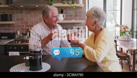 Discutant couple senior dans la chemise à carreaux et le pull tenant des tasses à café à l'île avec la presse française Banque D'Images