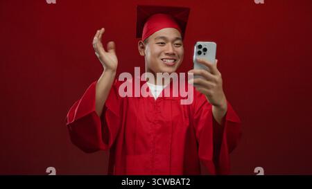 Jeune homme chinois étudiant en robe de graduation rouge faisant un appel vidéo sur smartphone au-dessus d'un mur rouge isolé célébrant la réussite scolaire avec une joyf Banque D'Images