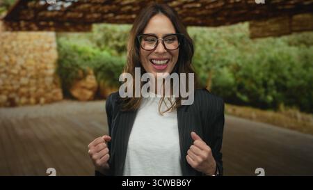 Jeune femme hispanique souriant en toute confiance dans un parc en plein air portant des lunettes et un costume rayé, exsudant le bonheur et le succès dans une envi naturelle Banque D'Images