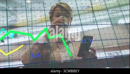 Femme d'affaires dans Blazer tenant la tablette et la tasse à café au bureau, en regardant les graphiques linéaires sur la grille de verre Banque D'Images