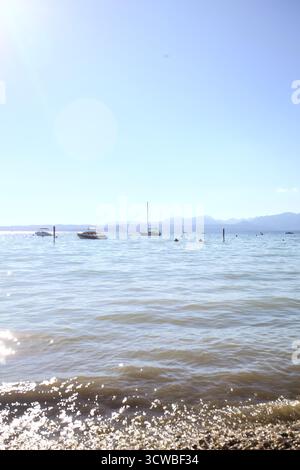 Plage de rochers avec des roseaux au bord du lac et des bateaux flottant au loin par une journée ensoleillée Banque D'Images