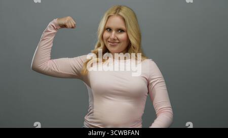 Femme fléchissant les muscles en toute confiance sur un fond gris lisse, portant une chemise rose décontractée, incarnant la force, l'autonomisation, la forme physique et la positiv Banque D'Images