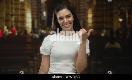 Femme hispanique jeune pointe pouce à côté à l'intérieur du bâtiment de l'église, souriant et debout par des bancs en bois sous la lumière intérieure chaude ; joie accueillante. Banque D'Images