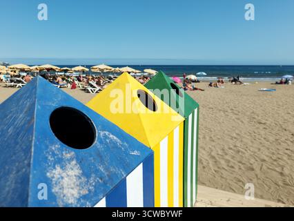 Trois grands bacs de recyclage à rayures colorées avec des ouvertures circulaires se dressent sur une plage de sable de Gran Canaria, entourés de chaises longues et de parasols. Banque D'Images