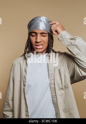 portrait d'un jeune homme aux cheveux tressés exprime confusion et frustration tout en se grattant la tête sur fond beige Banque D'Images