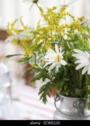 Les marguerites blanches et les fleurs sauvages jaunes créent une pièce maîtresse joyeuse dans un vase en métal rustique sur une table avec une nappe rayée rouge et blanche Banque D'Images