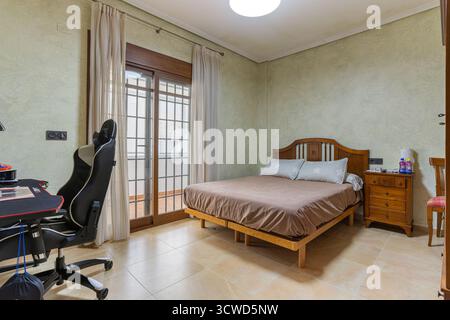 Cette chambre confortable et bien éclairée dispose d'un lit en bois avec literie marron, d'une table de chevet assortie et d'un bureau avec chaise de jeu. Verre du sol au plafond Banque D'Images