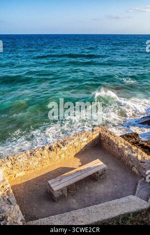 Banc en bois vide au bord de la mer turquoise avec des vagues éclaboussant sur la rive rocheuse sous le ciel bleu, scène estivale paisible symbolisant la détente, le voyage et le holid Banque D'Images