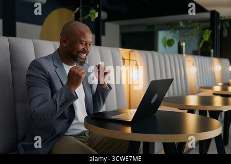 Homme d'affaires noir mûr joyeux serrant les poings tout en utilisant un ordinateur portable au bureau dans le salon de bureau Banque D'Images