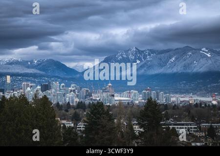 Des nuages sombres pendent au-dessus d'un horizon urbain avec des montagnes enneigées en arrière-plan. La scène capture une atmosphère hivernale sereine. Banque D'Images