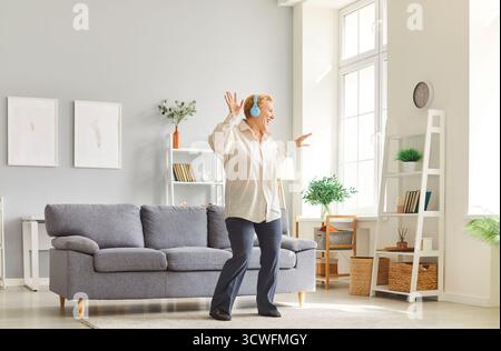 Femme mature joyeuse avec des cheveux blonds portant des écouteurs dansant dans le salon moderne lumineux. Banque D'Images