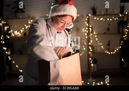 Jeune femme médecin dans des lunettes et chapeau de Père Noël sur le quart déballage de nourriture à emporter dans un sac en papier travaillant la nuit la veille de Noël à l'hôpital. Banque D'Images