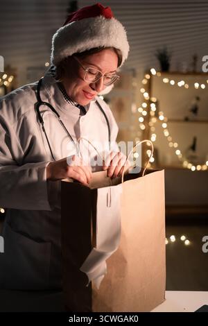 Jeune femme médecin dans des lunettes et chapeau de Père Noël sur le quart déballage de nourriture à emporter dans un sac en papier travaillant la nuit la veille de Noël à l'hôpital. Banque D'Images