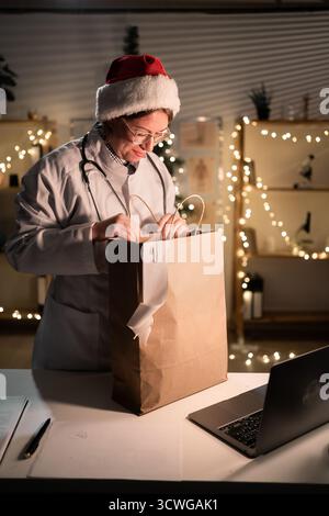 Jeune femme médecin dans des lunettes et chapeau de Père Noël sur le quart déballage de nourriture à emporter dans un sac en papier travaillant la nuit la veille de Noël à l'hôpital. Banque D'Images