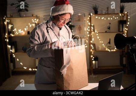 Jeune femme médecin dans des lunettes et chapeau de Père Noël sur le quart déballage de nourriture à emporter dans un sac en papier travaillant la nuit la veille de Noël à l'hôpital. Banque D'Images