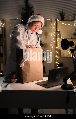 Jeune femme médecin dans des lunettes et chapeau de Père Noël sur le quart déballage de nourriture à emporter dans un sac en papier travaillant la nuit la veille de Noël à l'hôpital. Banque D'Images