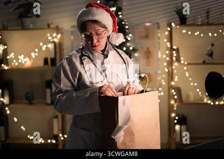 Jeune femme médecin dans des lunettes et chapeau de Père Noël sur le quart déballage de nourriture à emporter dans un sac en papier travaillant la nuit la veille de Noël à l'hôpital. Banque D'Images
