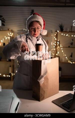 Jeune femme médecin dans des lunettes et chapeau de Père Noël sur le quart déballage de nourriture à emporter dans un sac en papier travaillant la nuit la veille de Noël à l'hôpital. Banque D'Images