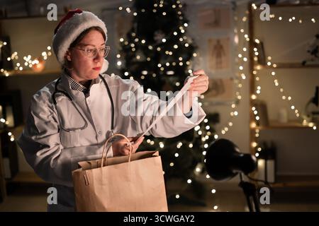 Jeune femme médecin dans des lunettes et chapeau de Père Noël sur le quart déballage de nourriture à emporter dans un sac en papier avec le prix travaillant la nuit la veille de Noël i Banque D'Images