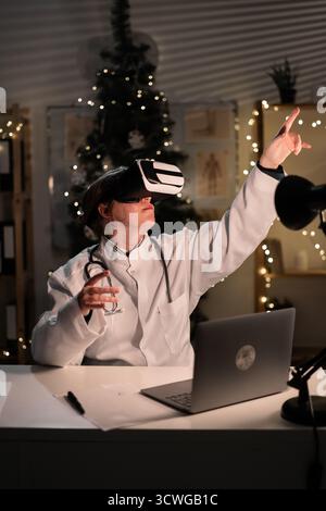 Jeune femme médecin porte un chapeau de Père Noël et des lunettes vr regardant la réalité metaverse sur le quart de travail la nuit la veille de Noël. Médecine, vacances, Banque D'Images