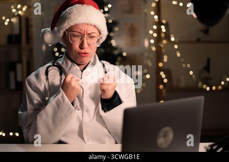 Jeune femme médecin porte le chapeau du Père Noël à l'aide d'un ordinateur portable tenant les poings sur le quart de travail la nuit la veille de Noël. Concept de médecine, vacances Banque D'Images